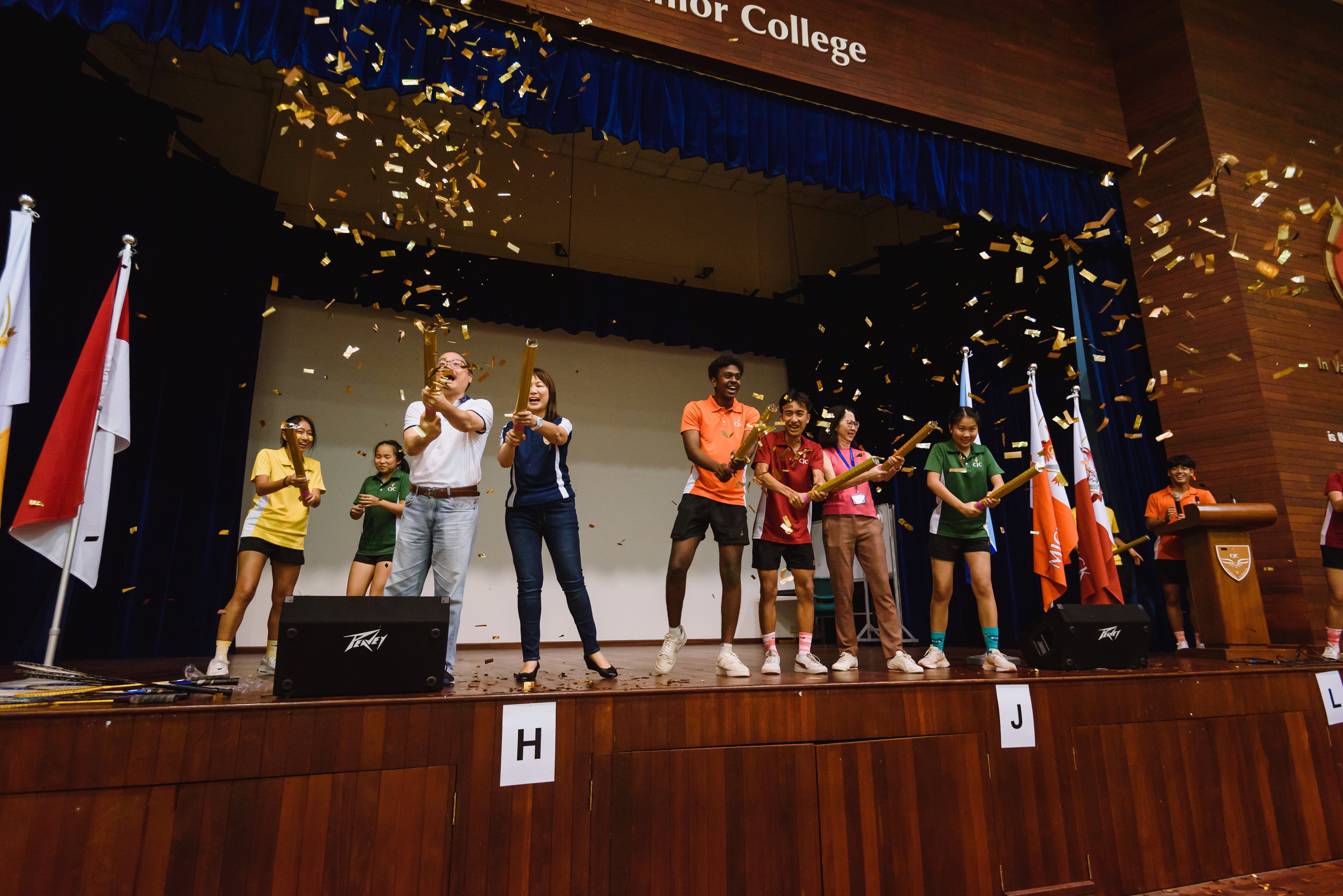 Group on stage pops confetti cannons as gold confetti rains down; flags at sides and a podium at right.